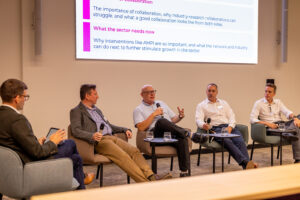 Five panellists seated on a stage during a conference discussion, with presentation slides displayed on a screen behind them.